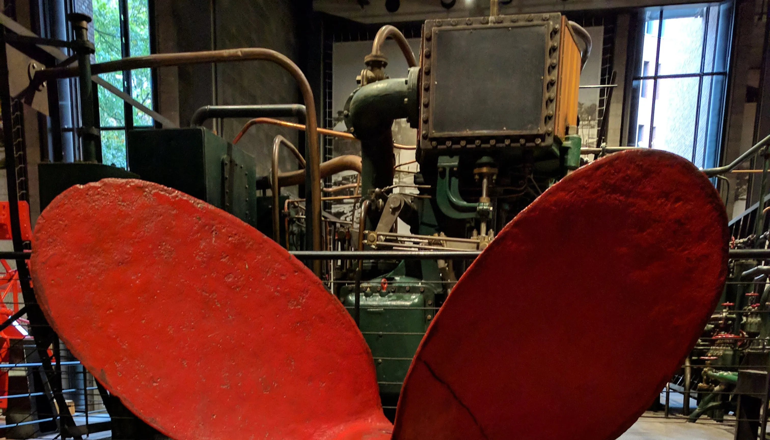 A bright red ship propeller in the foreground, steam pipes and machinery in the background.
