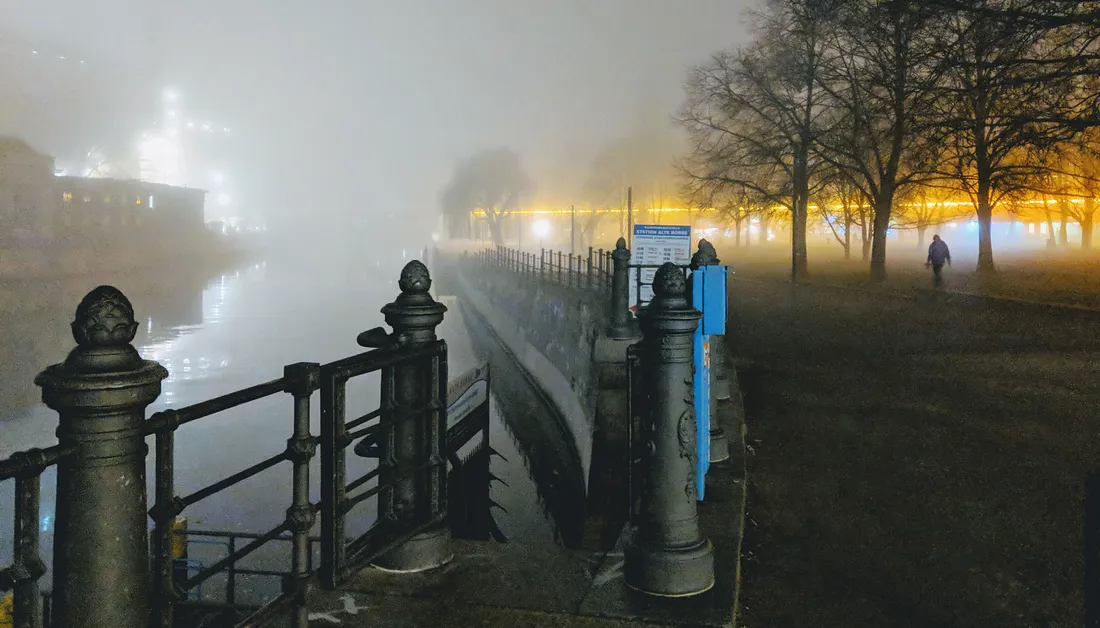 A dark evening in Berlin. The river borders a small city park park where a distant human figure walks towards a yellow light in the background.